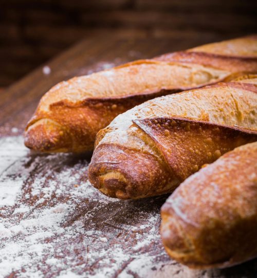 Close-up of fresh, golden-brown baguettes dusted with flour on a wooden table, showing texture and artisan quality.